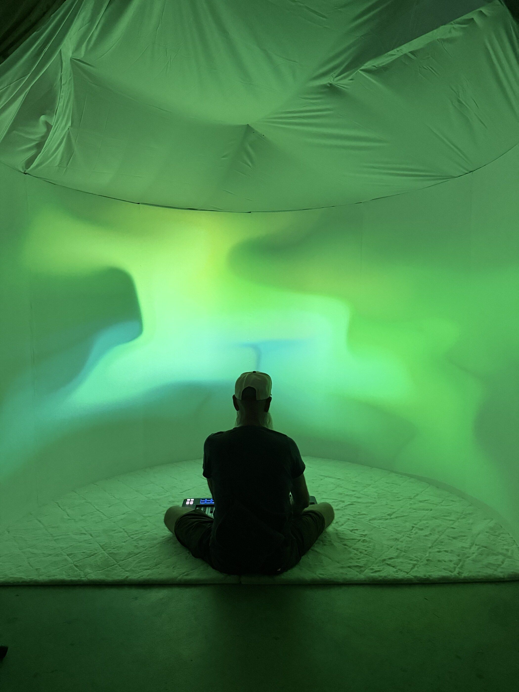 A visitor sitting alone in the installation, the curved wall around them washed green and blue.