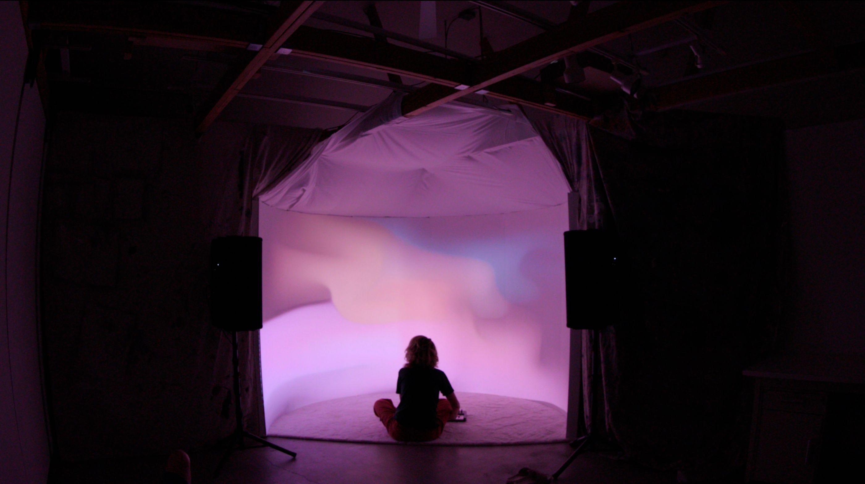 A visitor sitting alone in the installation while the wall runs violet and rose.